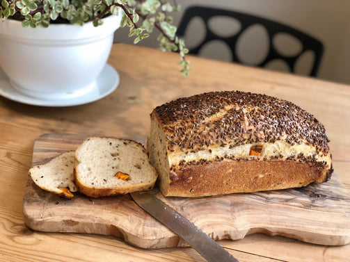 A loaf of artisan sourdough bread with Kumara and Flaxseed, cut into a slice, on a wooden board.